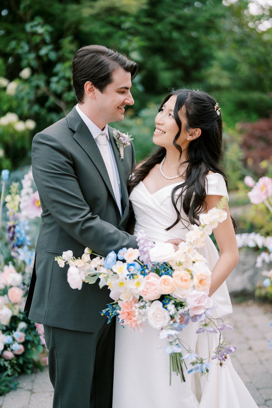 A happy couple in formal wedding attire stands in a garden setting, surrounded by lush greenery. The bride, holding a vibrant bouquet of pink, white, and blue flowers, gazes affectionately at the groom, capturing a romantic and joyful moment.