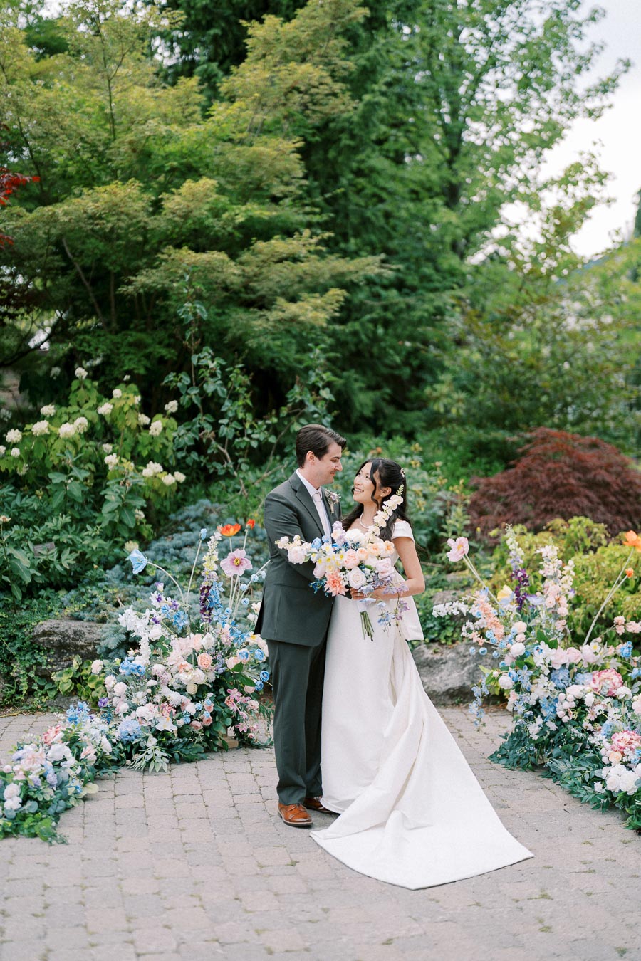 A bride and groom stand closely together in a lush garden setting, surrounded by colorful floral arrangements. The bride is wearing a white wedding gown and holding a bouquet of pastel flowers. The groom is in a dark suit, smiling at the bride. The backdrop includes vibrant greenery and blooming flowers, creating a romantic and serene atmosphere.