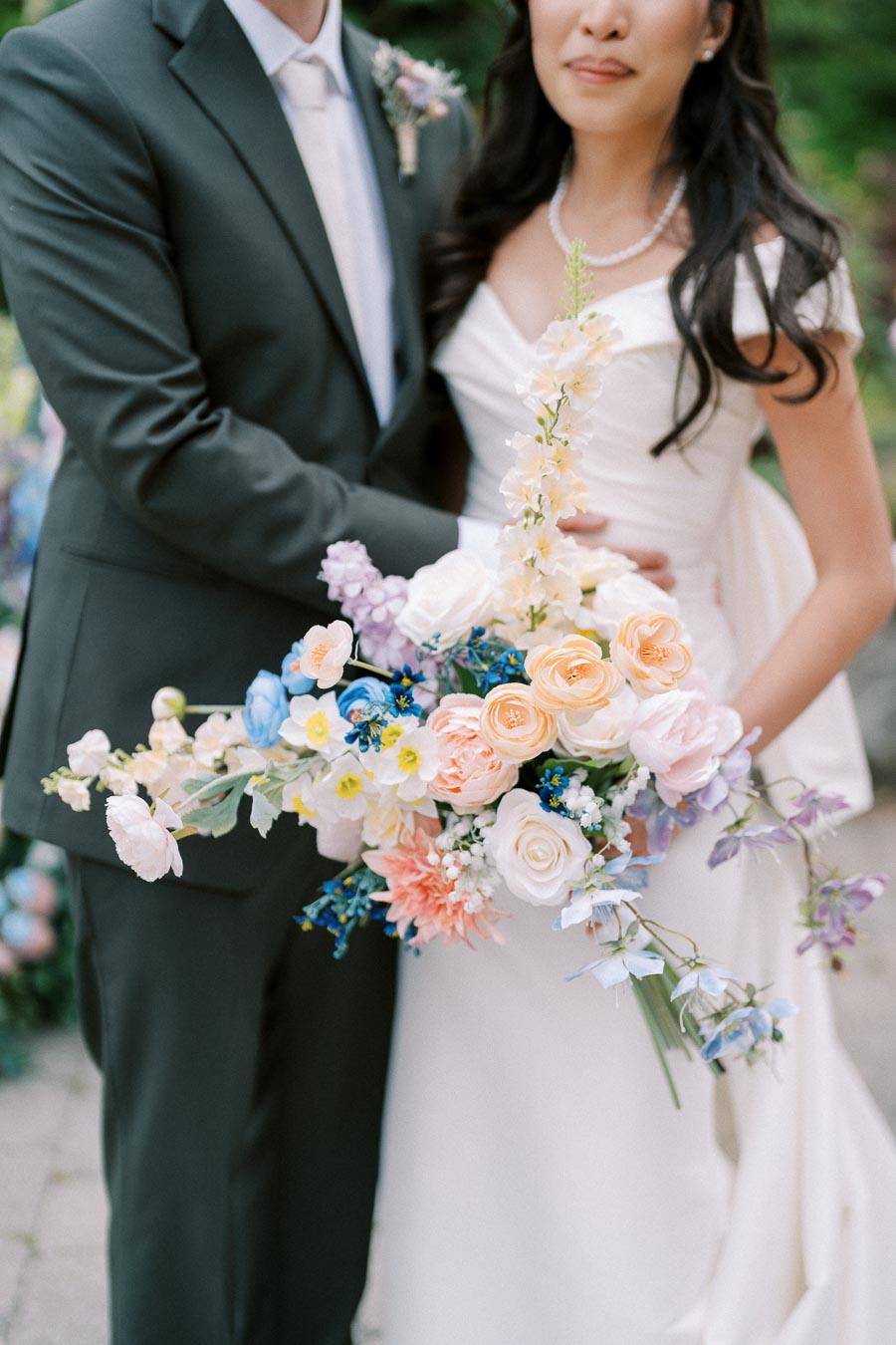 Bride in an elegant white gown holding a colorful bouquet of roses and wildflowers, standing beside a groom in a dark suit, capturing a romantic wedding moment outdoors.