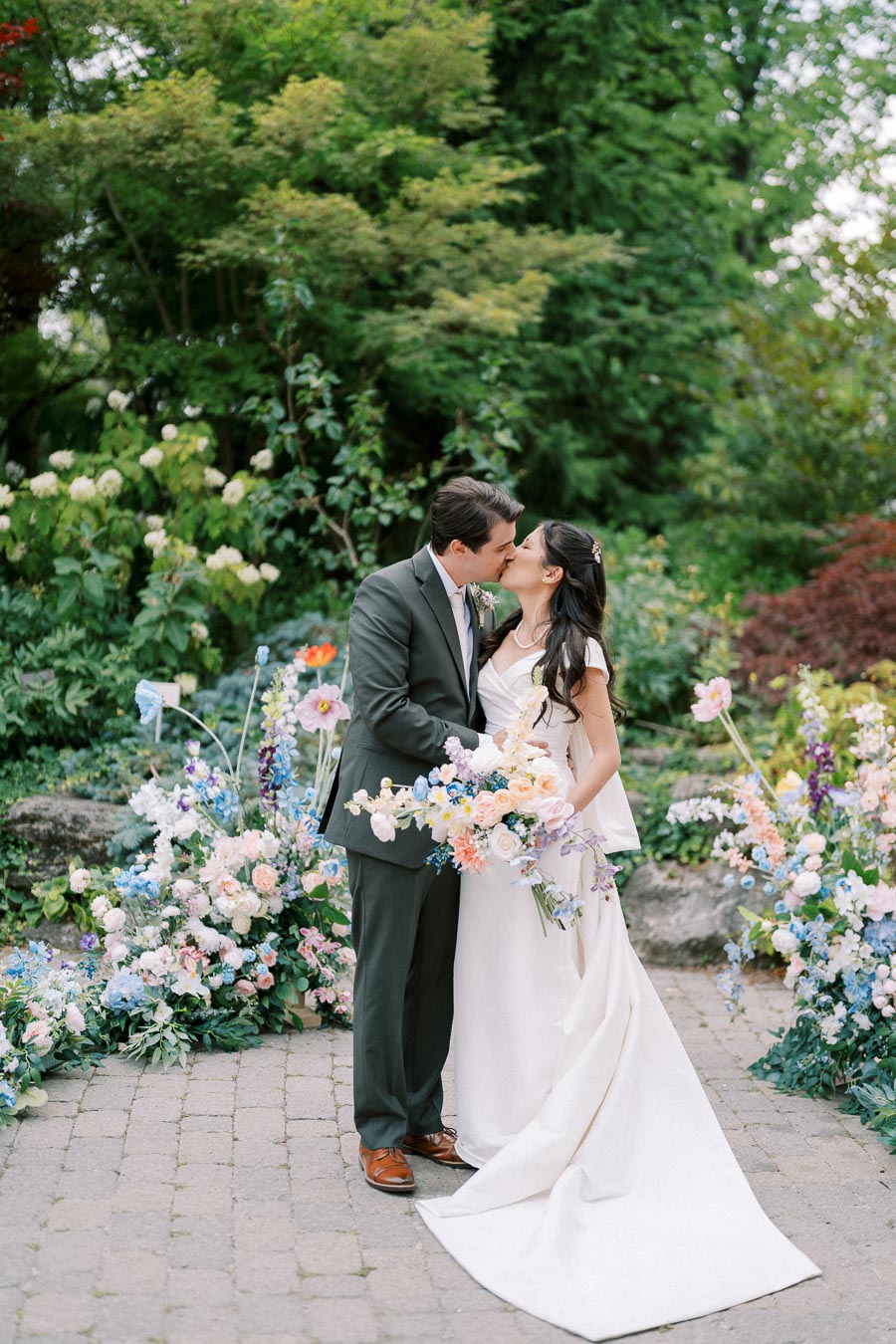 Bride and groom kissing in a lush garden, surrounded by colorful floral arrangements, during their outdoor wedding ceremony.