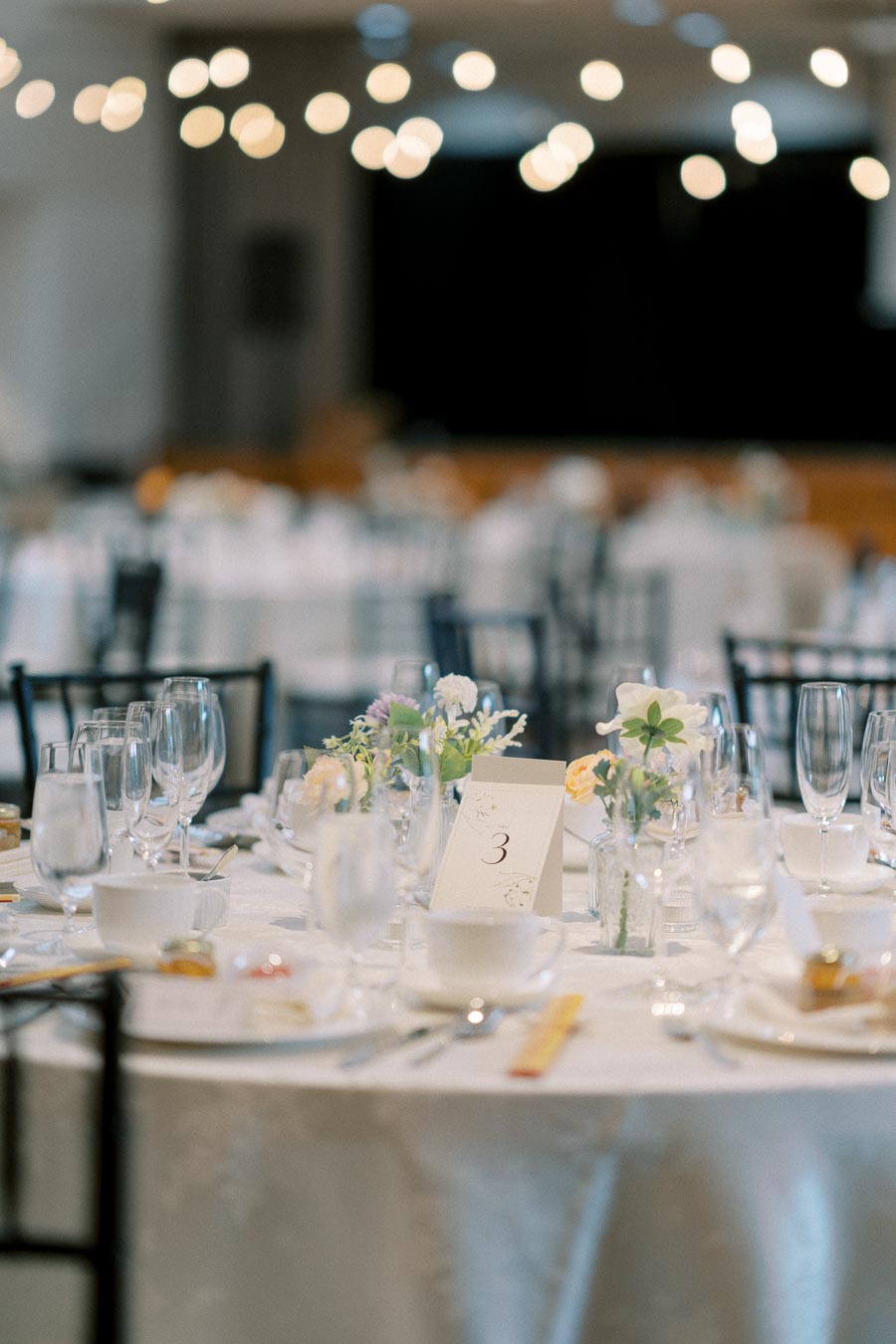 Elegant wedding reception table setting with floral centerpieces, glassware, and table number three, softly lit with string lights in the background.