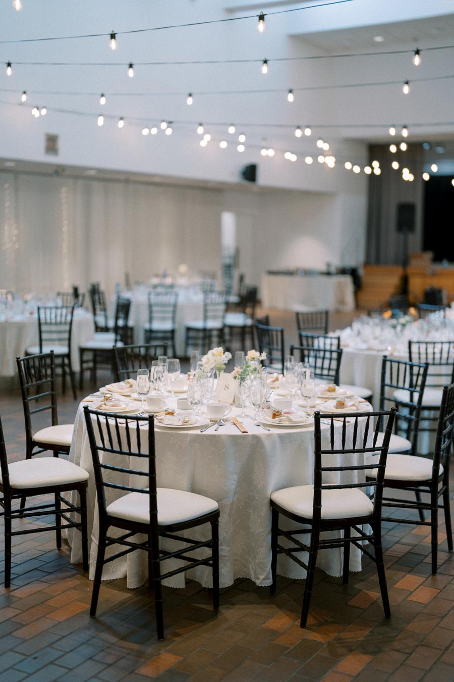 Elegant wedding reception setup with round tables draped in white tablecloths, adorned with floral centerpieces and surrounded by dark wooden chairs. String lights hang overhead, creating a warm and inviting atmosphere for guests.