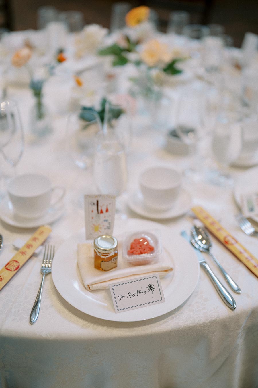 Elegant wedding table setting with a white plate, small jar of honey, and a container of pink candies on a white napkin. The table is adorned with floral decorations and includes chopsticks, a fork, and a card with calligraphy.