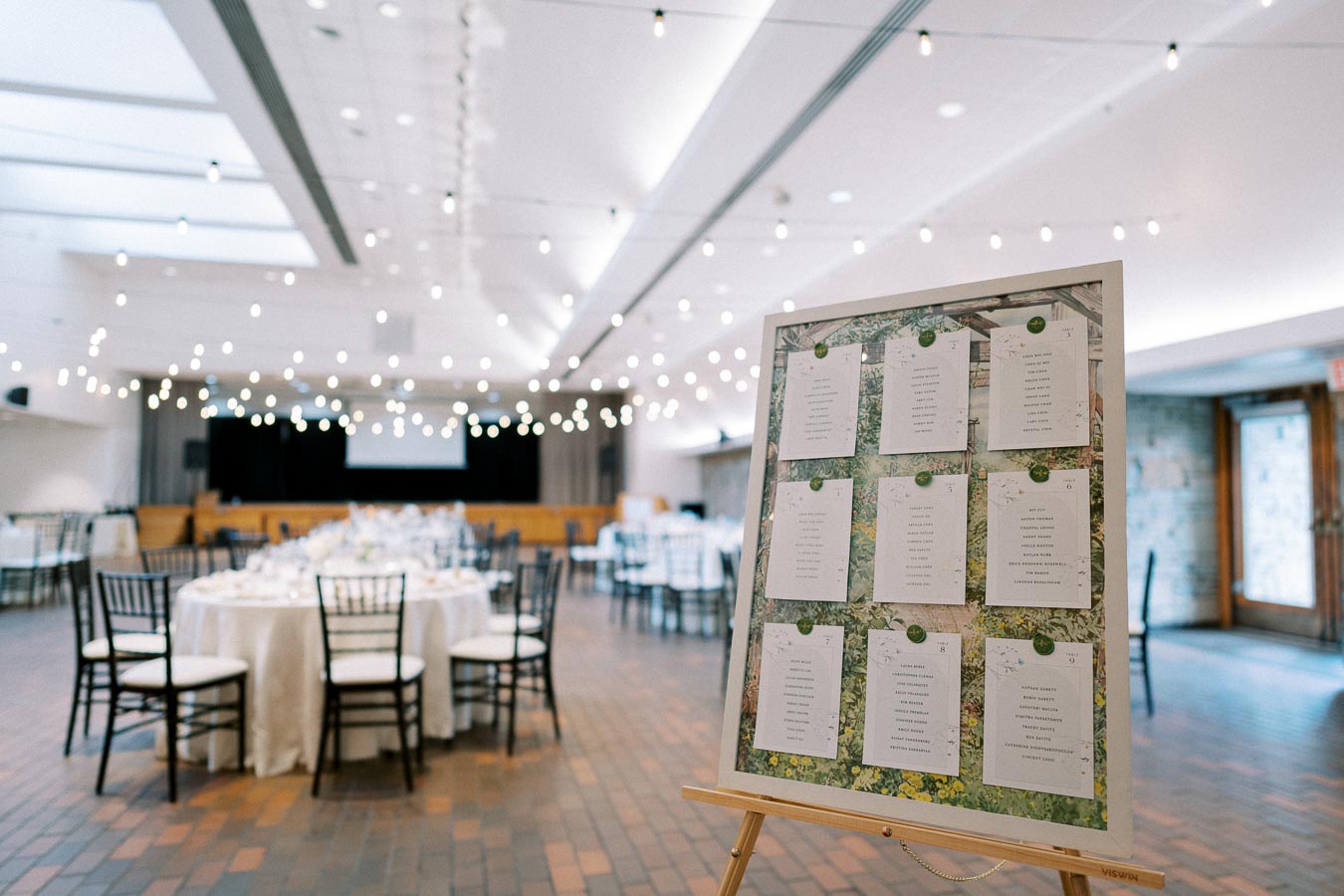 Elegant wedding reception hall with string lights and round tables, featuring a prominent seating chart on an easel in the foreground.