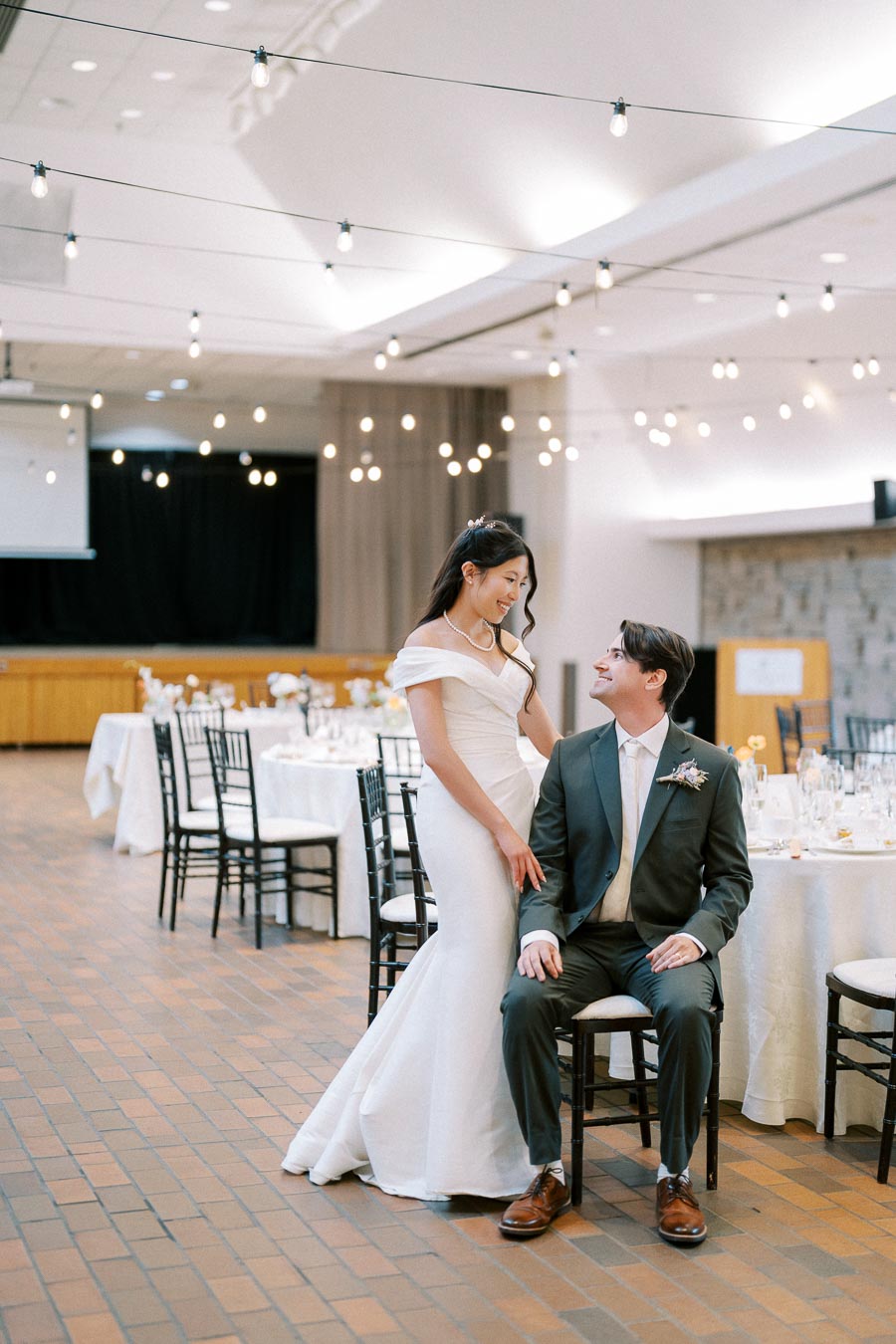 A bride in an elegant white gown and a groom in a dark suit, smiling at each other in a beautifully decorated wedding reception hall with string lights and round dining tables.