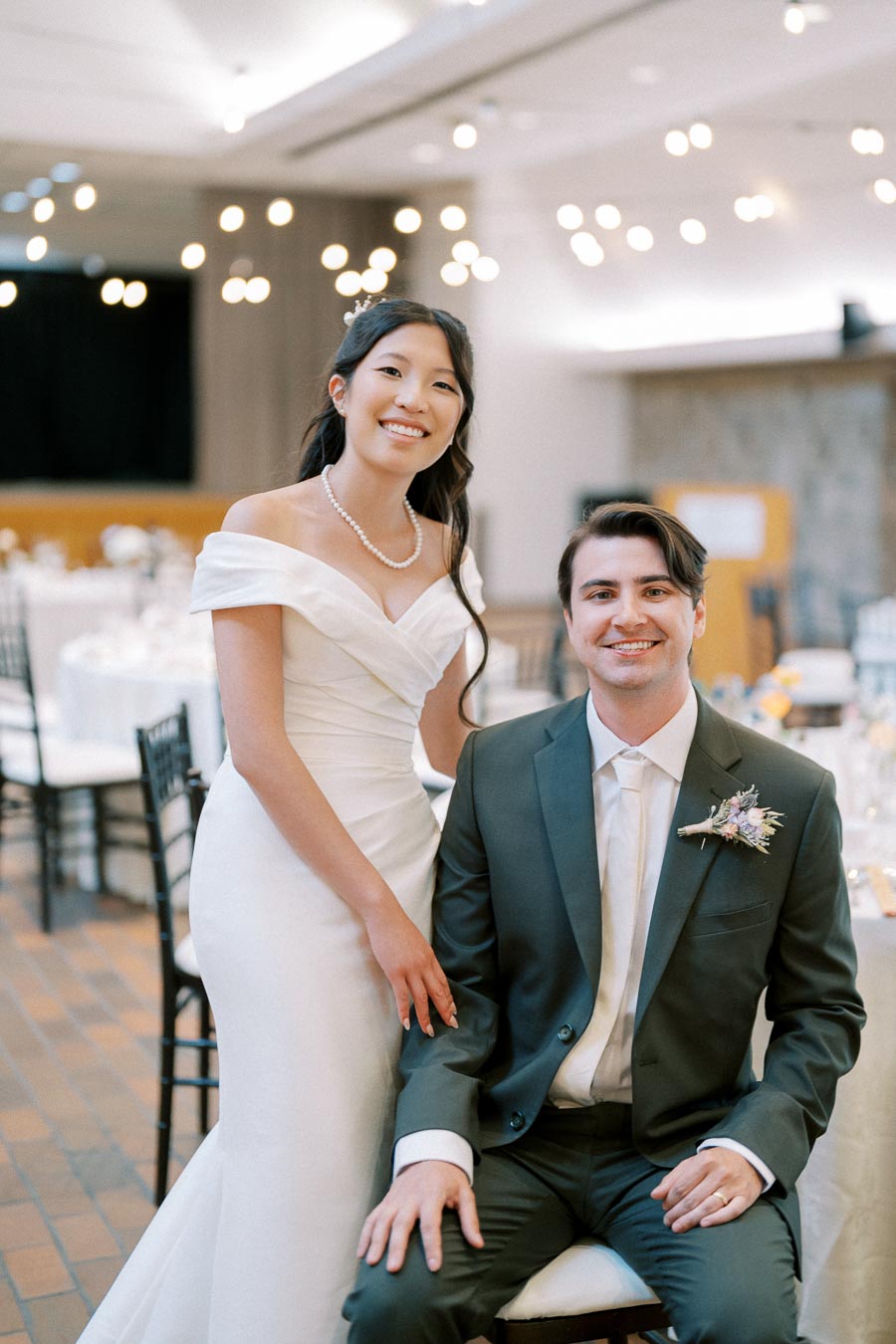 Bride in an elegant white gown standing beside groom in a dark suit at a wedding reception with soft lighting and round tables in the background.