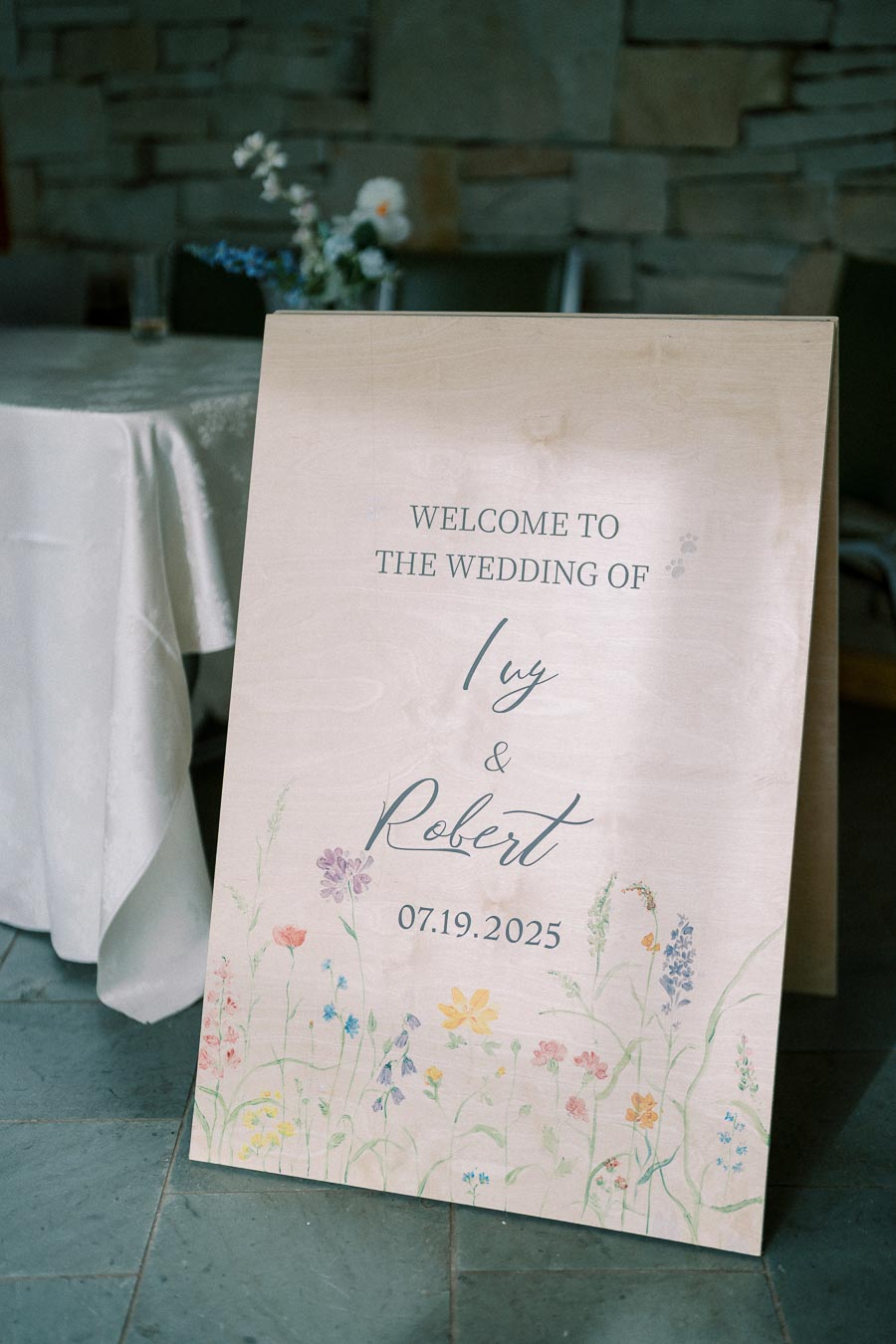 Wedding welcome sign for Ivy and Robert on July 19, 2025, featuring floral designs at the bottom, set against a stone-wall and table background.