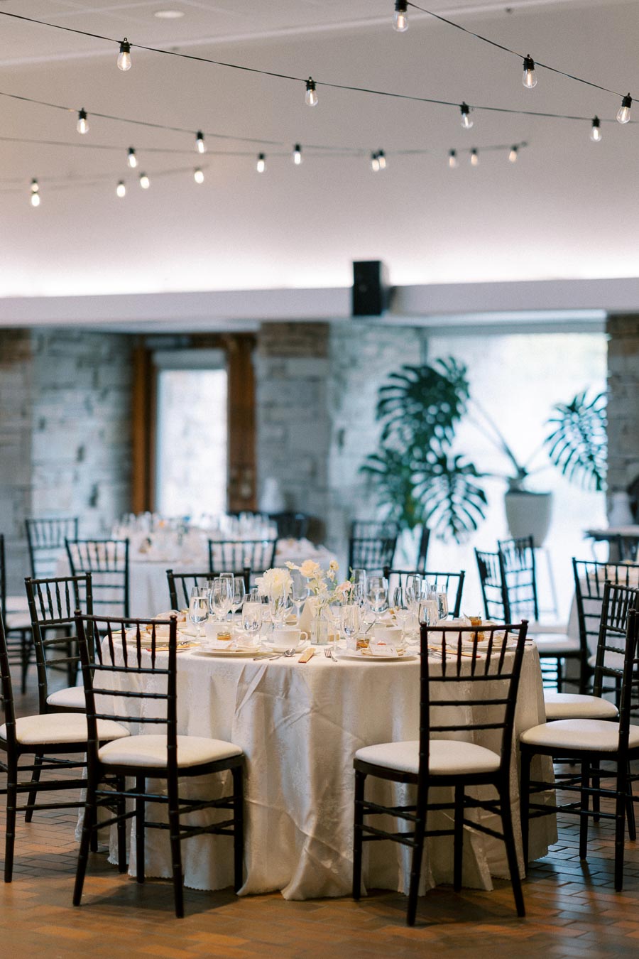 Elegant wedding reception setup with round tables covered in white tablecloths, surrounded by black chairs, adorned with glassware, white floral centerpieces, and twinkling string lights overhead.