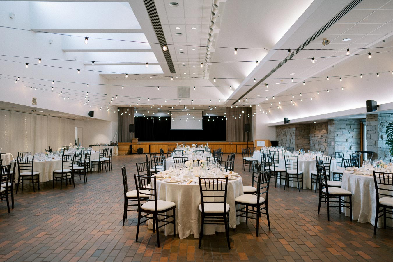 Elegant banquet hall set up for an event, featuring round tables with white tablecloths, black chairs, and overhead string lights creating an inviting ambiance.