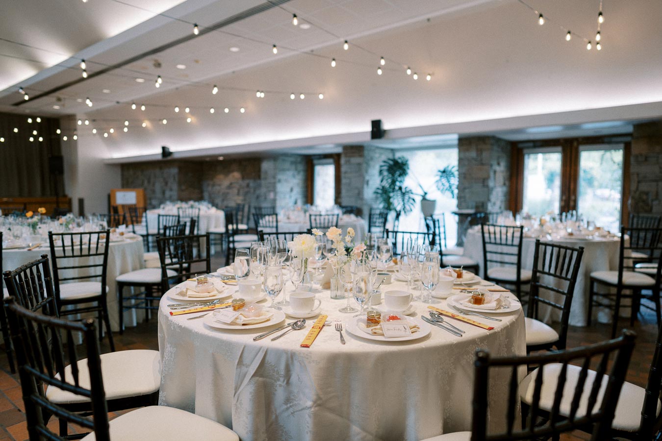 Elegant wedding reception setup in a spacious venue, featuring round tables dressed with white linens, neatly arranged silverware, and glassware. Soft lighting and decorative string lights create a romantic ambiance.