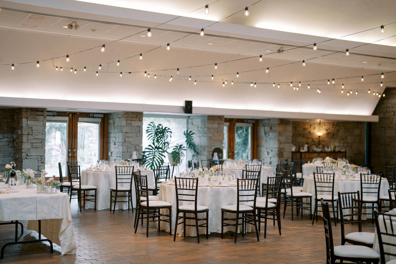 Elegant banquet hall with round tables set for an event, featuring white tablecloths, dark wooden chairs, and ambient string lights hanging from the ceiling, creating a sophisticated and inviting atmosphere.