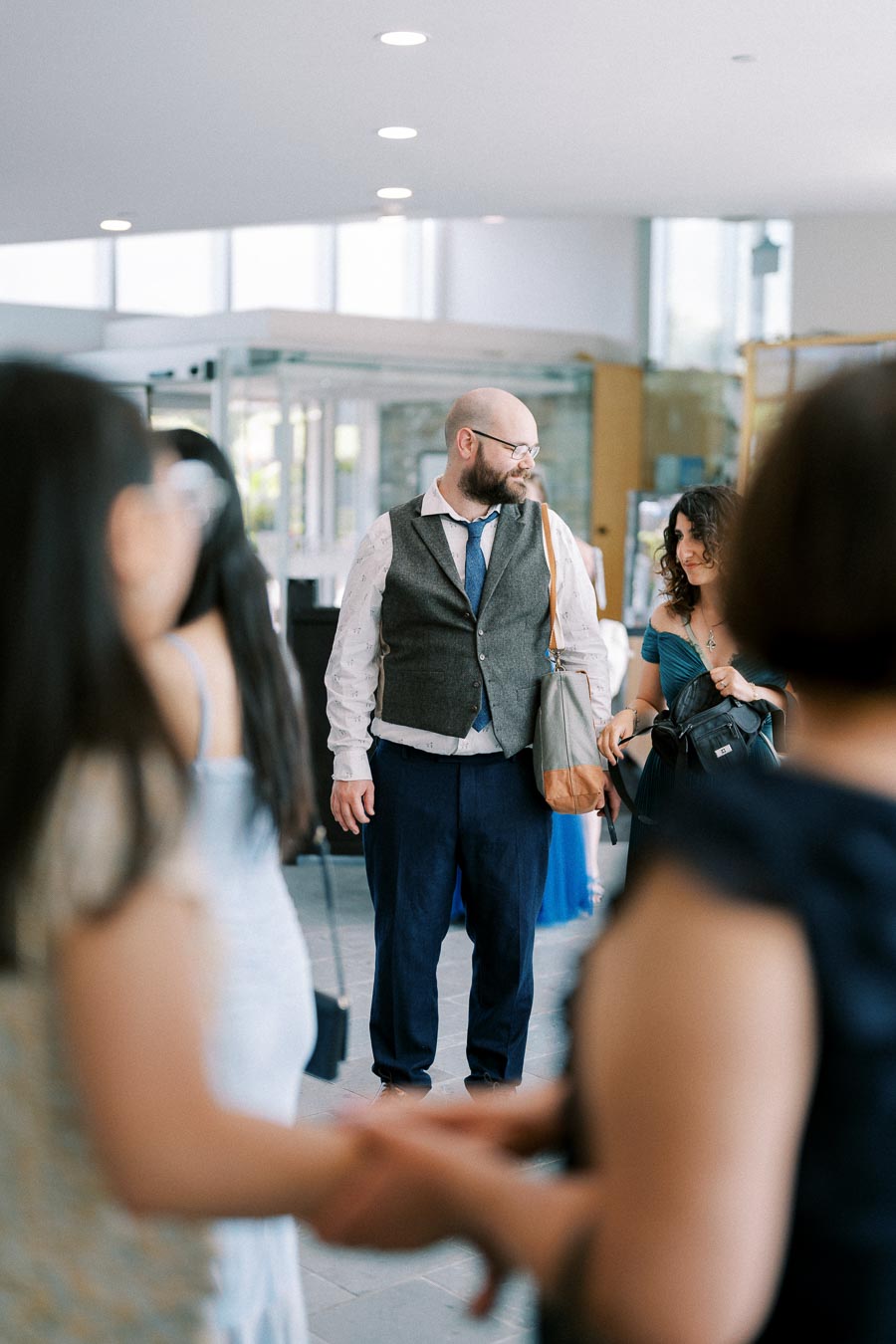 A group of formally dressed individuals interacting at a modern indoor event, featuring a man with a beard wearing a vest and carrying a shoulder bag, engaging in conversation with a woman in a blue dress. Bright, well-lit venue with glass walls in the background.