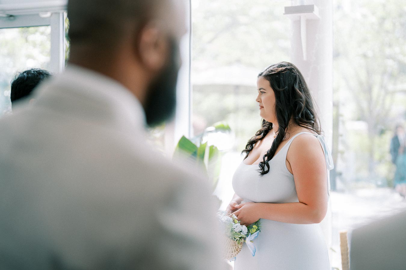 A woman in a light blue dress holding a small bouquet stands indoors, gazing thoughtfully towards a window filled with natural light while other people are partially visible in the foreground.