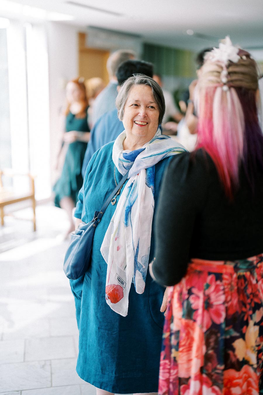Smiling older woman in blue dress and scarf talking to a woman with pink hair and floral skirt in a bright, indoor social setting.