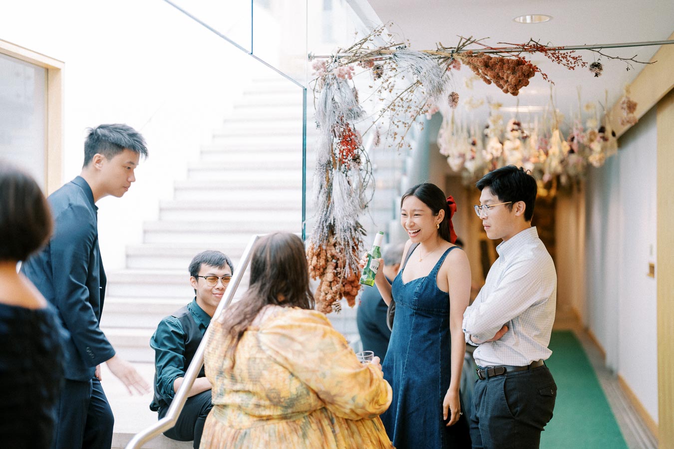 A group of people socializing in a well-lit hallway decorated with hanging dried flowers, with one person holding a beverage and others engaged in conversation.