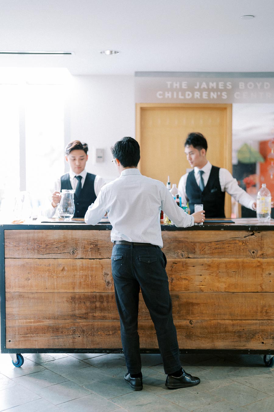 Man in white dress shirt and black pants at a wooden bar counter with two bartenders in vests preparing drinks, in a bright, indoor setting.