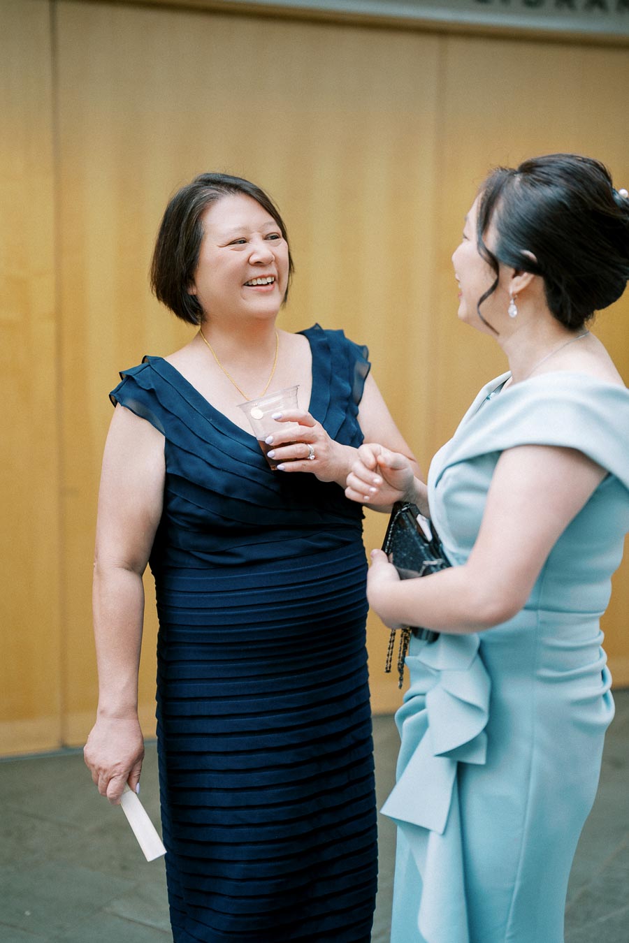Two women in elegant dresses having a joyful conversation at a social event, with one holding a drink. The setting appears formal, indicating a celebration or gathering.
