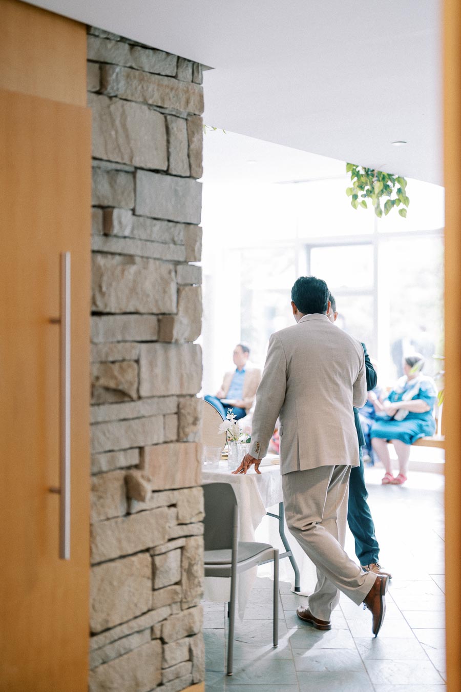 Man in light-colored suit standing near a stone wall in a bright, modern interior space with people seated in the background.