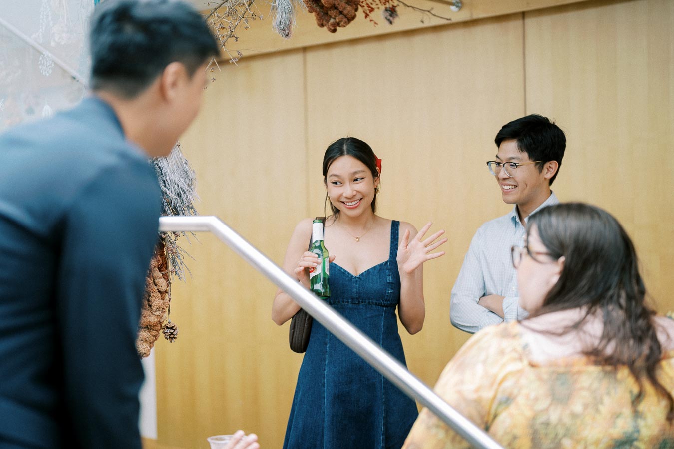A group of people enjoying a casual social gathering indoors, with a woman in a denim dress holding a beer bottle and smiling, surrounded by others engaged in conversation.
