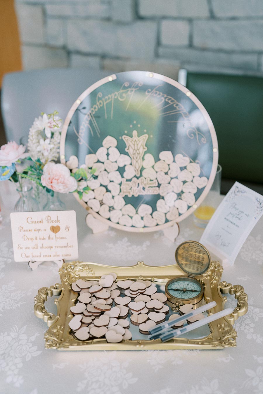 Elegant wedding guest book display with heart-shaped tokens on a decorative golden tray. A circular frame with inscriptions is filled with these tokens, accompanied by flowers and pens, set on a lace-covered table.