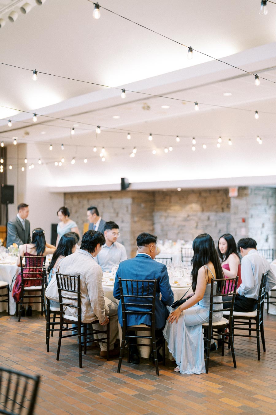 Guests sitting at a decorated table in a stylish indoor wedding reception with string lights and a brick wall background.