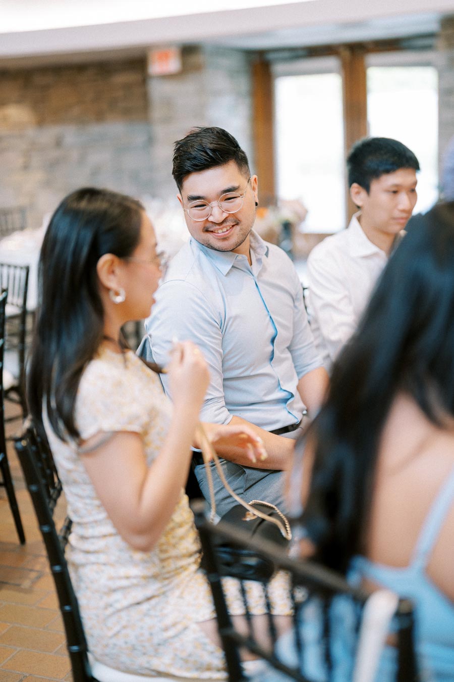 A group of people at an indoor event, sitting and conversing, with a focus on a man in a light blue shirt smiling at a woman in a floral dress.