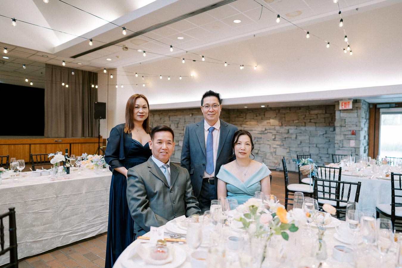 Elegant group portrait at a formal event, featuring four adults dressed in suits and gowns, seated around a beautifully decorated table with floral arrangements in a well-lit venue.