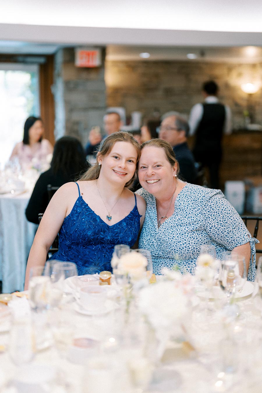A mother and daughter smiling at a wedding reception, sitting at a beautifully set table with floral centerpieces, surrounded by other guests in formal attire.
