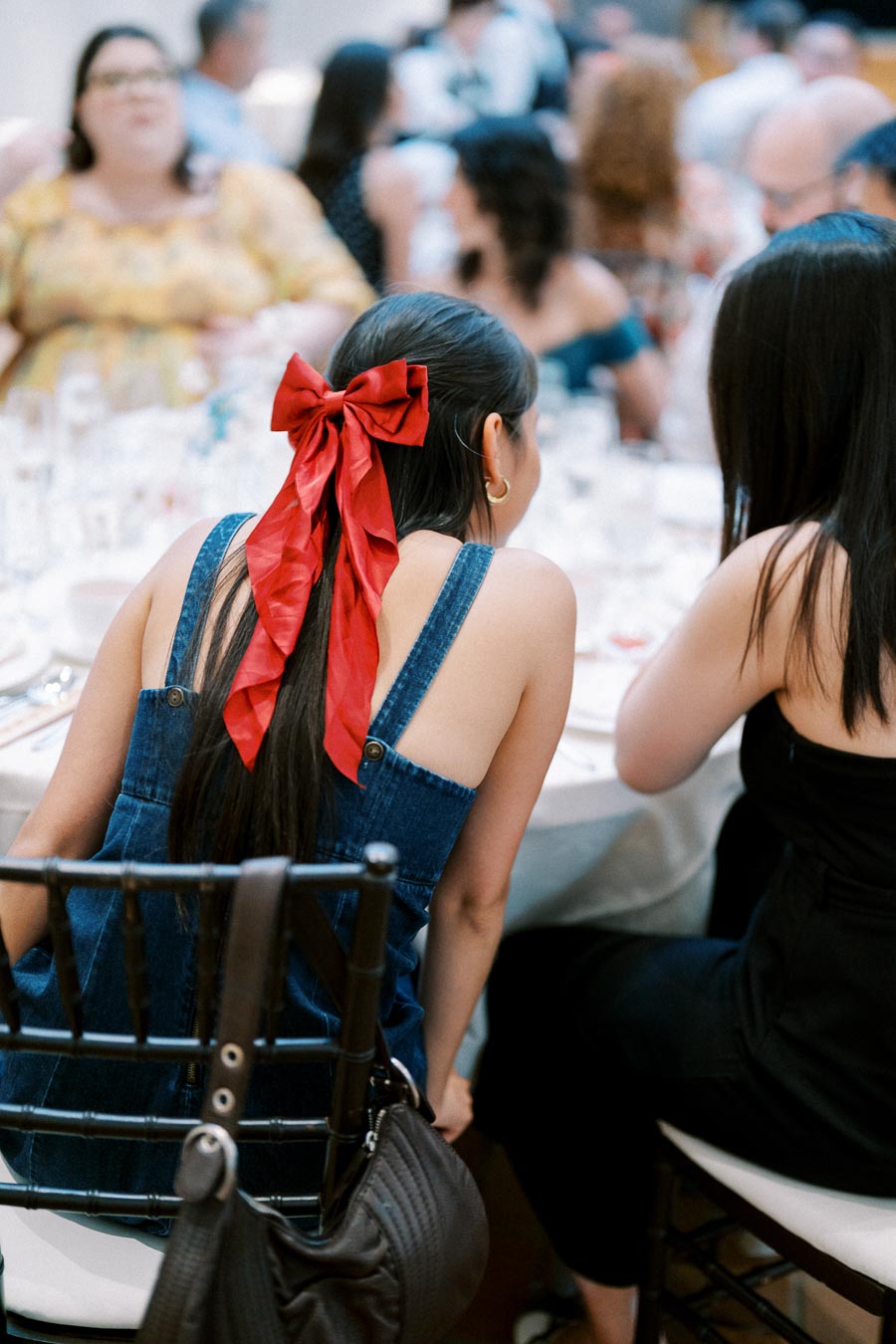 Young woman with long dark hair adorned with a large red bow, wearing a denim top, sitting at a table during a lively social gathering.