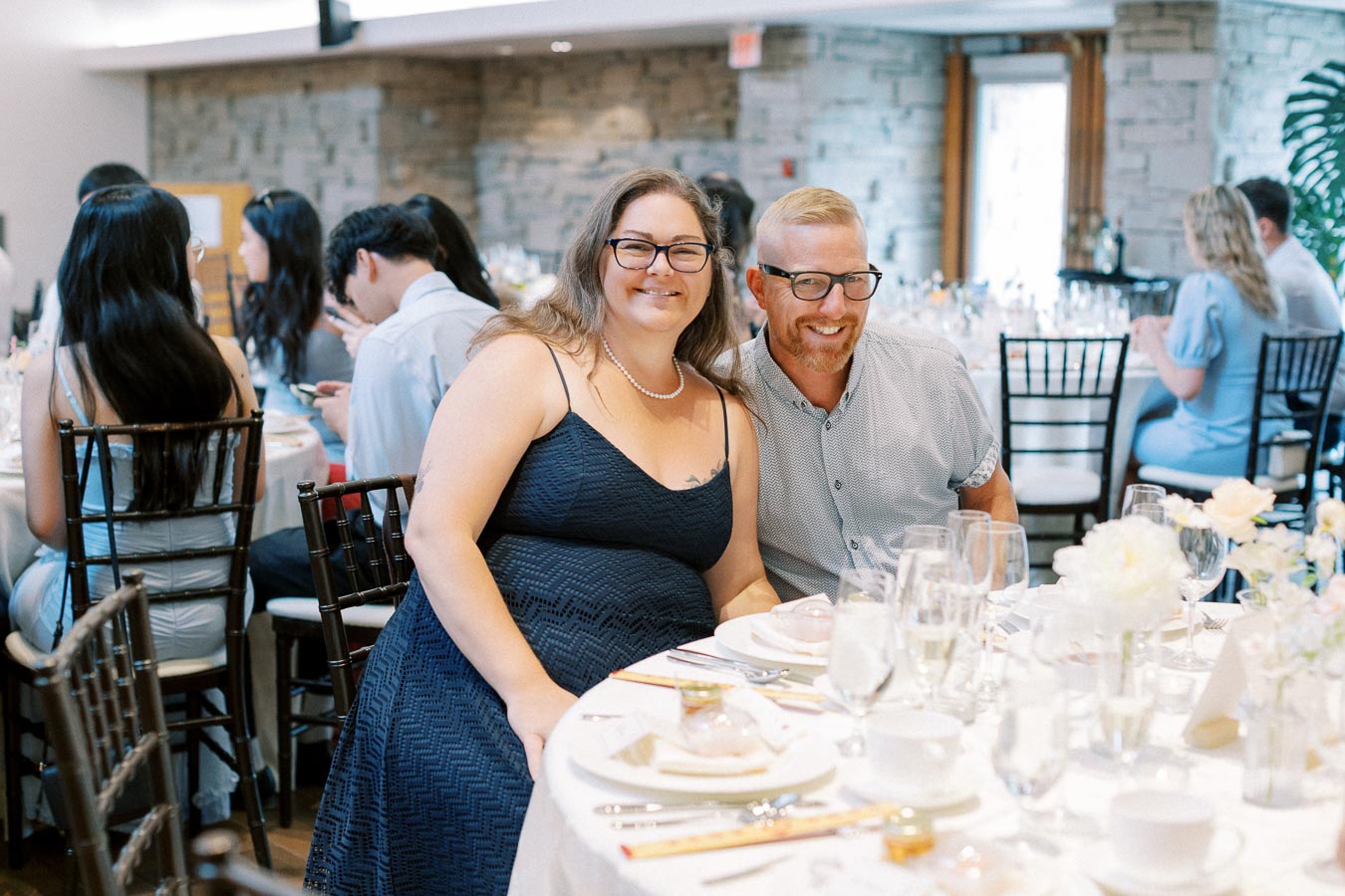 Smiling couple seated at a wedding reception table with elegant decor and guests in the background.