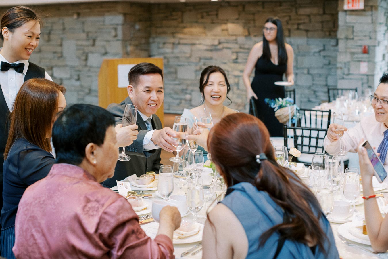 A joyful group of people celebrating at a formal event, raising glasses in a toast around a beautifully set dining table, with a stone wall and a server in the background.