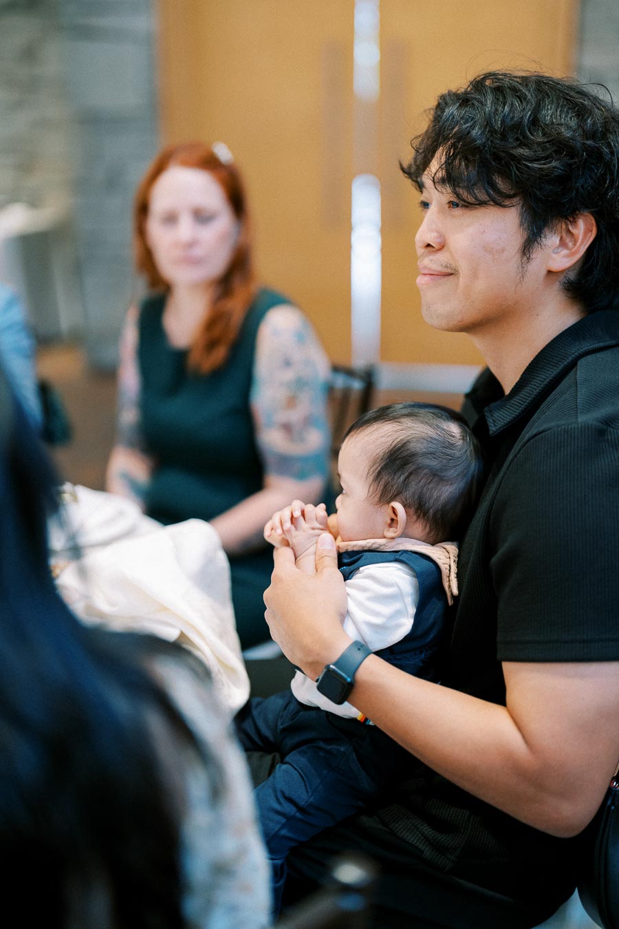 Man holding a baby at a gathering, with a woman in the background.