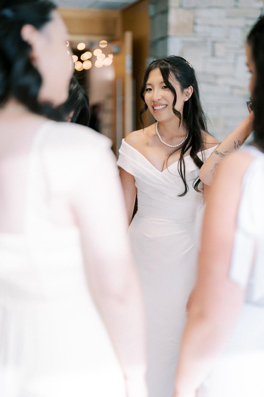 A bride in an elegant off-the-shoulder white wedding dress smiling indoors, surrounded by bridesmaids, creating a joyful wedding preparation moment.