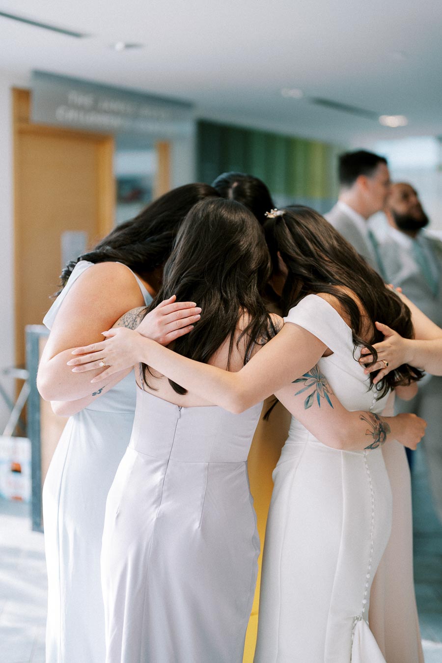 A group of bridesmaids in elegant dresses share a heartfelt hug before a wedding ceremony in a bright indoor venue.