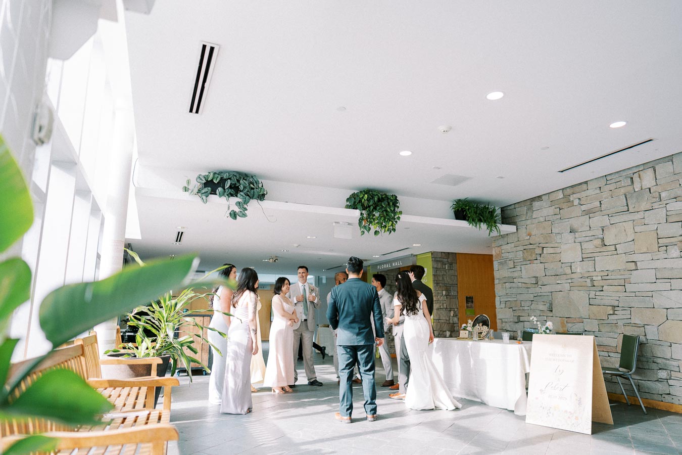 Group of elegantly dressed guests mingling in a bright, indoor event space with stone walls and lush green plants.