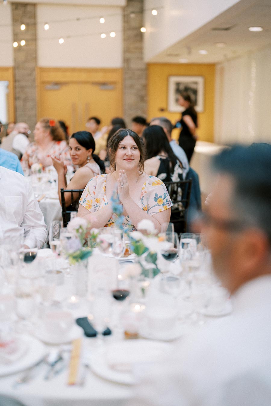Wedding reception with guests seated around a decorated table, including a woman clapping and others engaging in conversation, in a warmly lit venue.