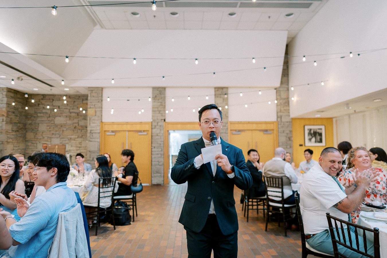 A man in a suit giving a speech at a formal event reception in a decorated hall, with guests seated at tables, listening and clapping.