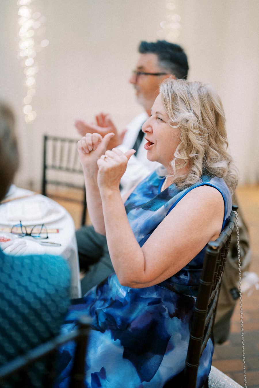 Elegant woman with curly blonde hair in a blue dress expressing joy at a formal event, while seated at a decorated table indoors.
