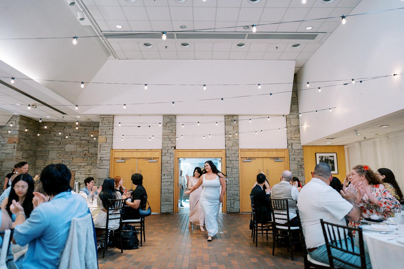 A joyous wedding reception scene featuring elegantly dressed guests seated at tables, with two women in white dresses entering the venue under string lights, capturing a celebratory and festive atmosphere.