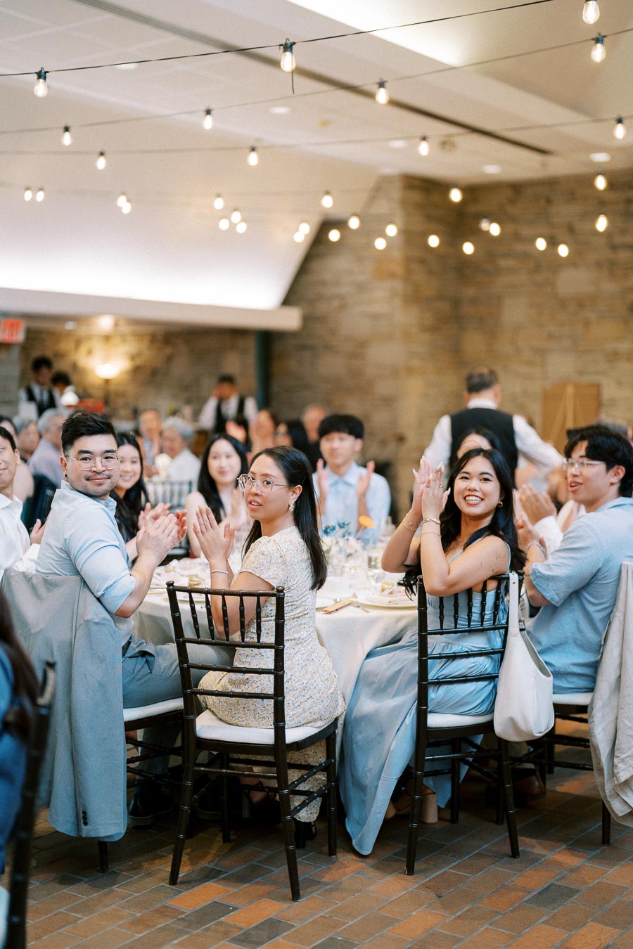 Guests seated at a round table, clapping and celebrating at an elegant indoor event with string lights and stone wall decor.