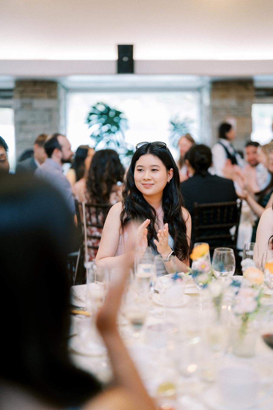 A woman with long dark hair clapping at a formal event, surrounded by people seated at a table with wine glasses and floral decorations.