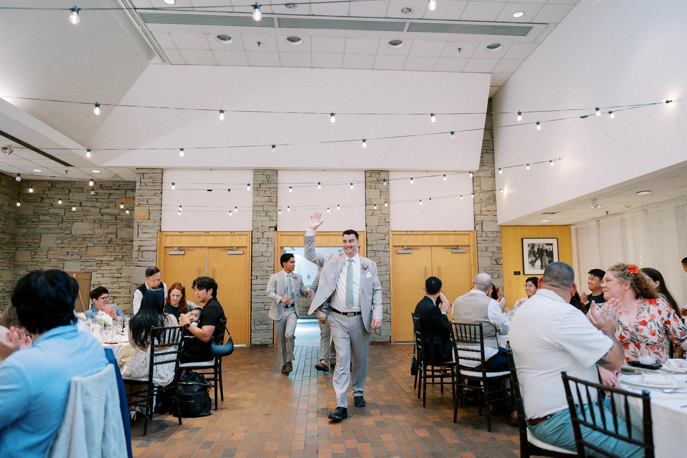 Wedding reception in a decorated banquet hall with guests seated at tables, groom walking in smiling and waving, ambient string lights overhead.