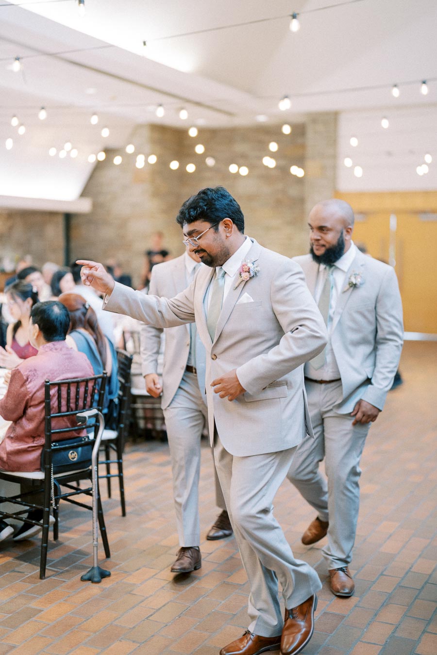 A group of groomsmen in matching light gray suits walk with enthusiasm at a wedding reception. The scene is set indoors with string lights creating a festive atmosphere as seated guests watch.