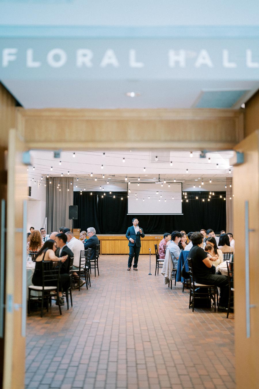 Indoor event at Floral Hall featuring guests seated at round tables and a speaker on stage, with string lights creating a warm ambiance.