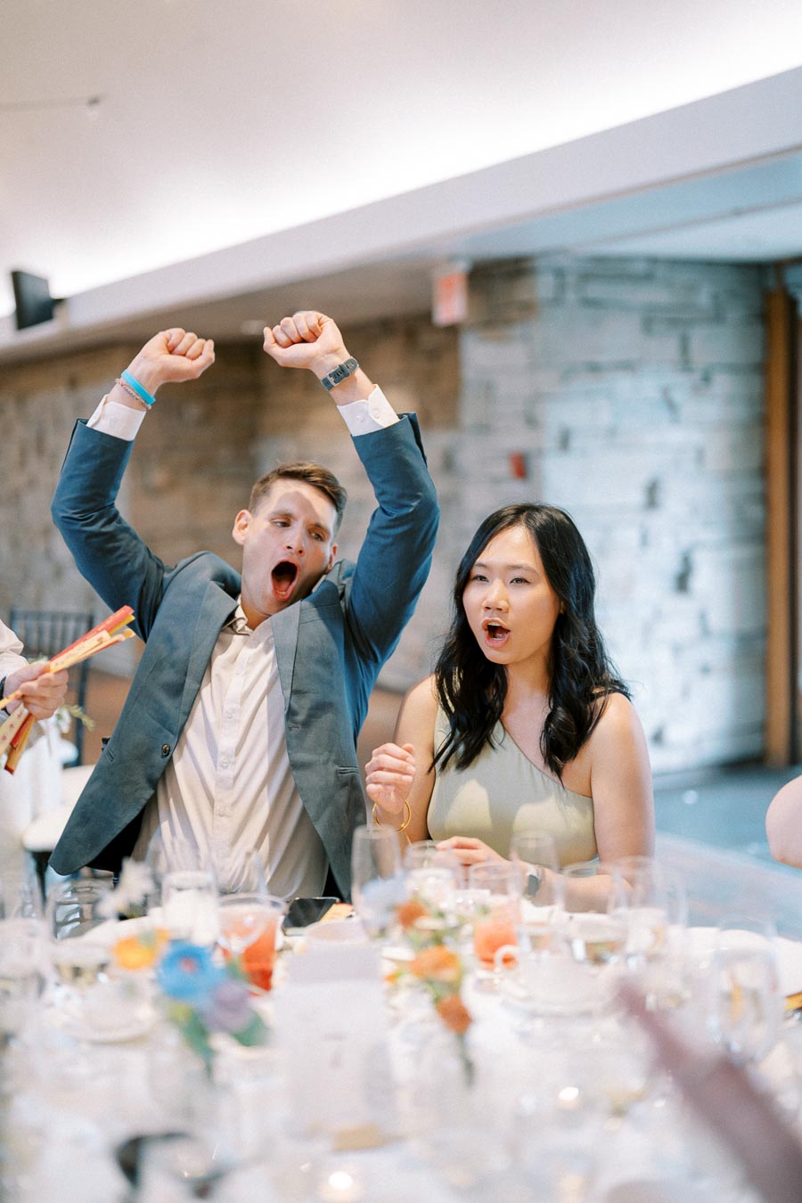 Excited people celebrating at a wedding reception, with a man in a blue suit raising his arms and a woman in a dress looking surprised, sitting at a table adorned with floral arrangements and glassware in a modern banquet hall.