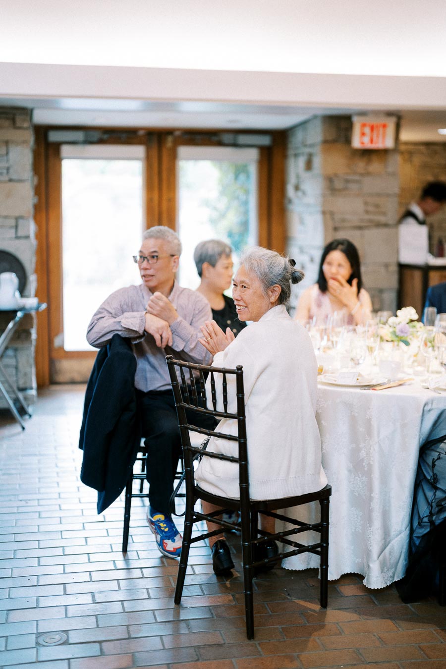 Elderly guests enjoying a conversation and clapping at a formal indoor event, seated around a decorated table in a warmly lit room with brick walls.