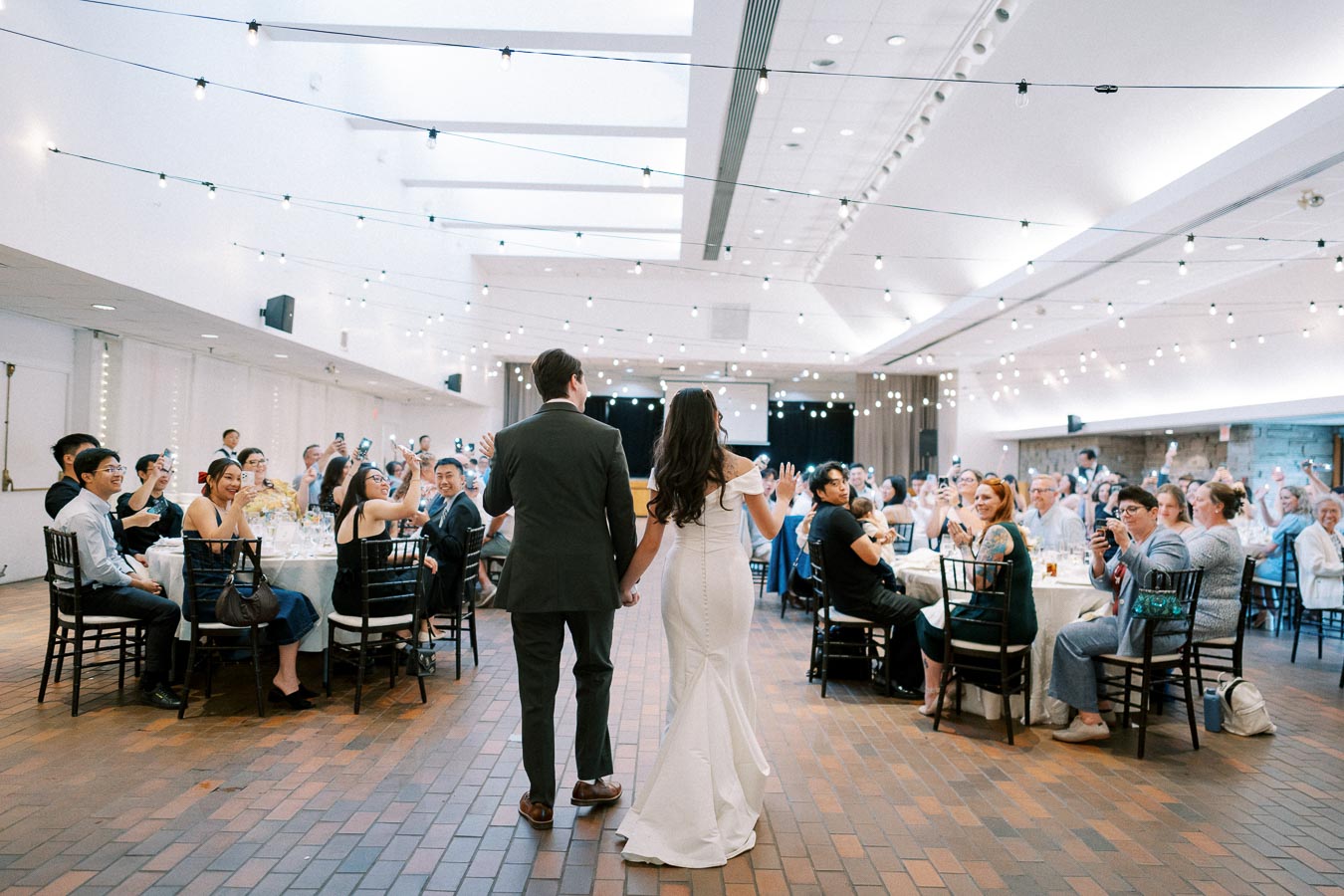 A newlywed couple holding hands enters a beautifully decorated reception hall filled with guests seated at round tables. String lights illuminate the room, creating a festive atmosphere as attendees cheer and take photos.