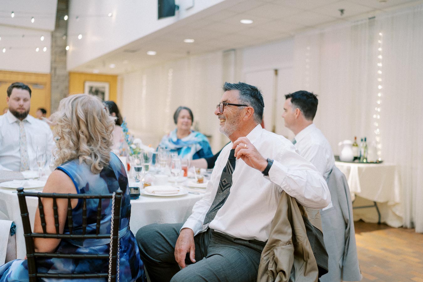 Elegant wedding reception with guests seated at a round table, adorned with white tablecloths and surrounded by soft lighting, creating a warm and festive atmosphere.