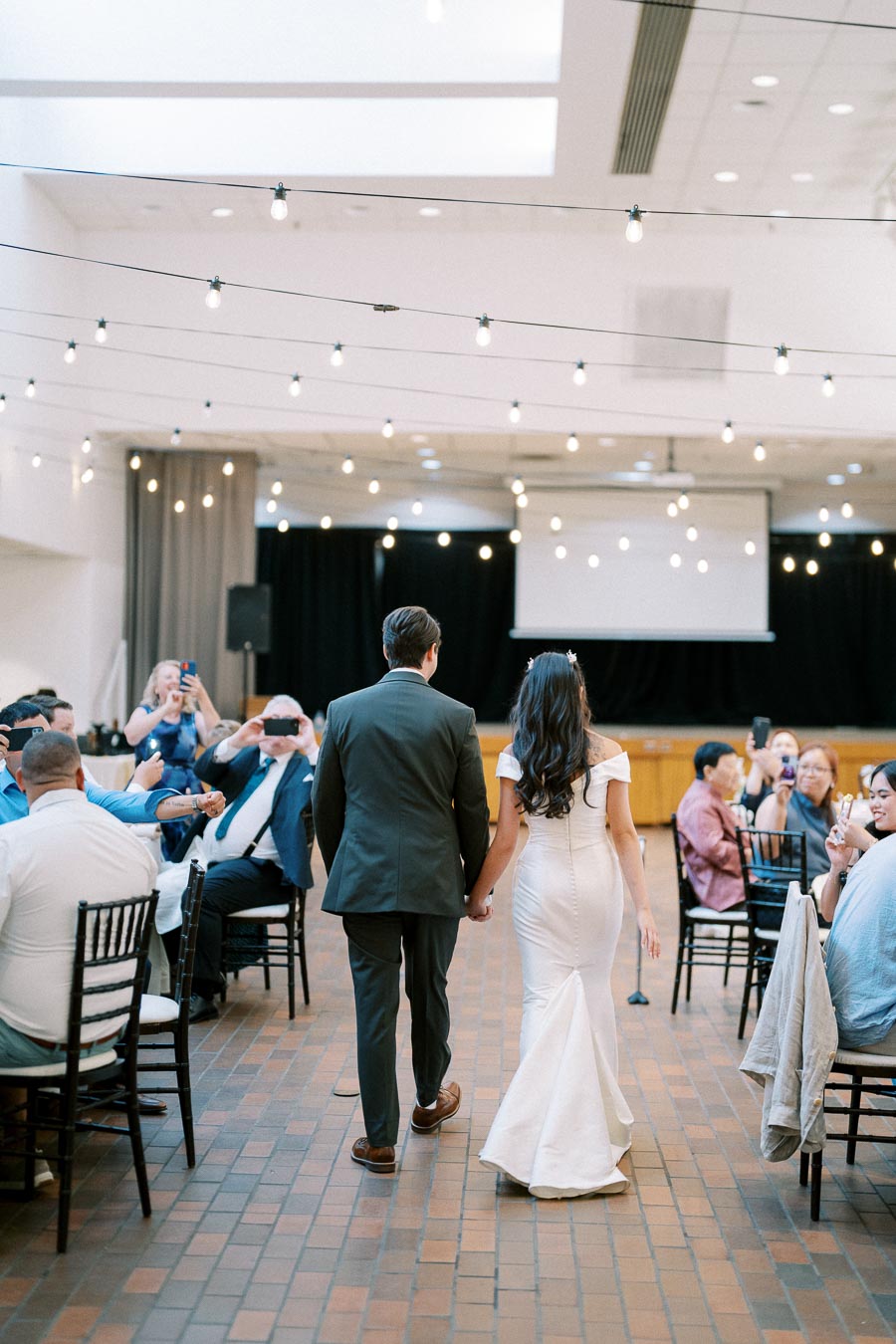 Wedding celebration with a bride in a white gown and groom in a suit holding hands as they walk through a decorated reception venue under string lights, surrounded by seated guests taking photos.