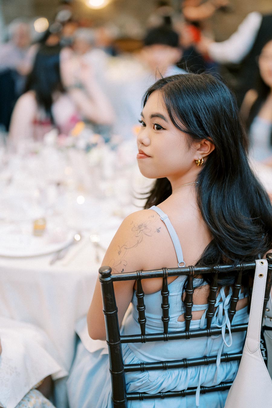 Young woman with long dark hair and floral tattoo, wearing a light blue dress, sits at an elegantly set dinner table, looking backwards with a thoughtful expression.